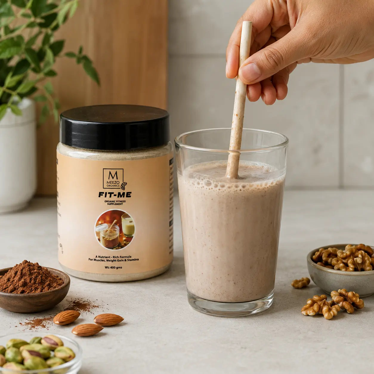A hand holds a wooden straw in a glass of creamy beige drink beside a jar labeled FIT-ME. The scene includes almonds, walnuts, pistachios, cocoa powder, and a plant on a light stone surface.